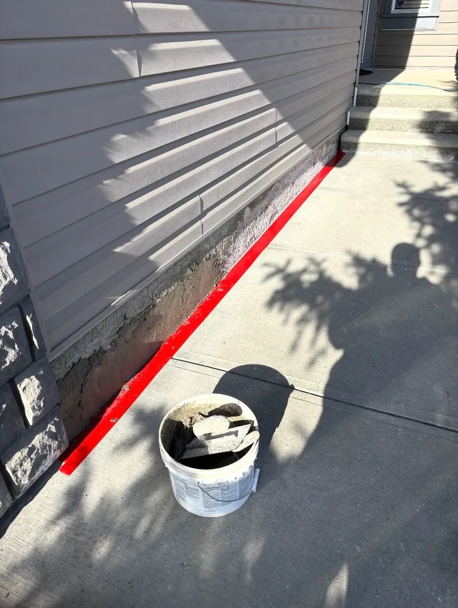 Calgary parging repair before — old parging chipped away, bare concrete exposed, red tape masking off the work area, fresh mortar bucket on site