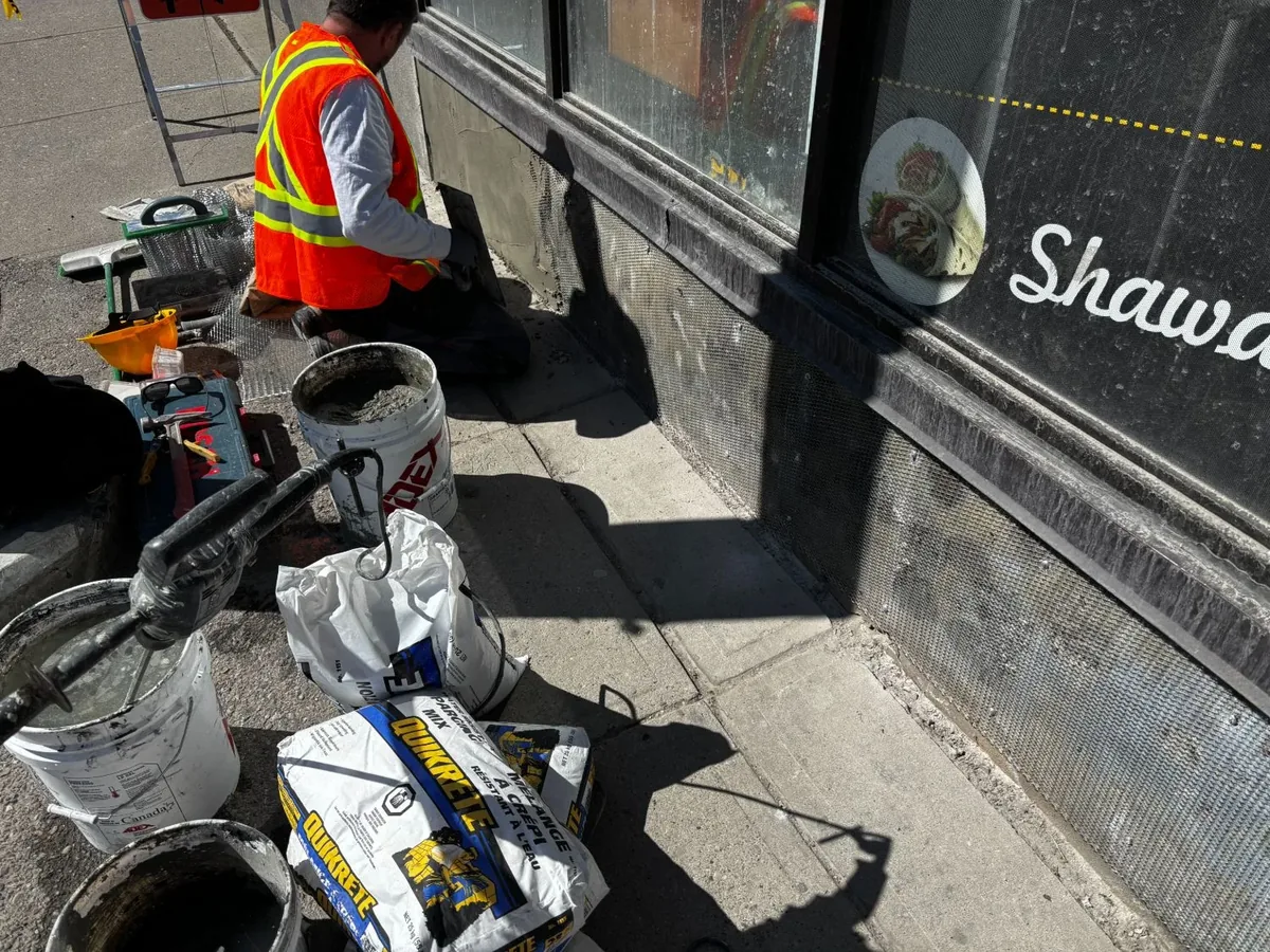 Calgary parging crew applying polymer-modified cement on a commercial storefront base — mesh-lath visible on the wall, Quikrete polymer-modified mix bags and fresh mortar bucket on site