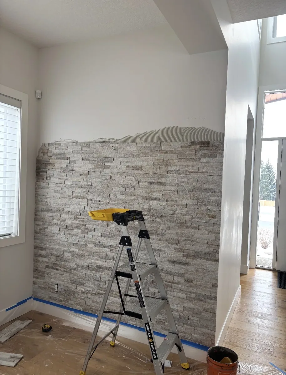 Calgary interior stone accent wall mid-installation — thinset applied at top, lower portion of stacked ledgestone in place, ladder and tools on drop cloth