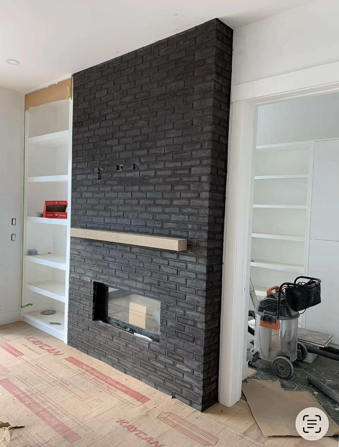 Calgary custom charcoal brick fireplace mid-build — floor-to-ceiling dark painted brick with rustic wood mantel and built-in shelving