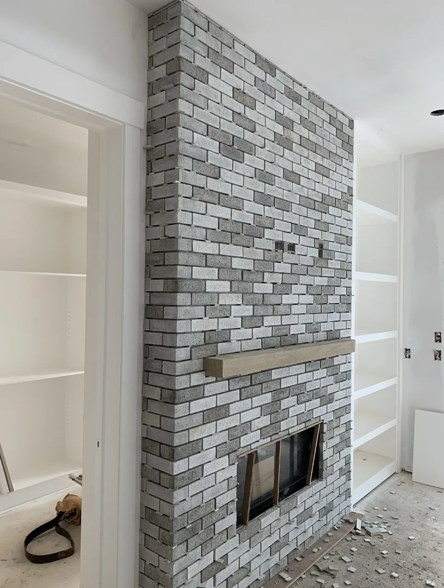 Calgary custom whitewash brick fireplace mid-build — floor-to-ceiling light-grey brick with wood mantel and built-in shelving