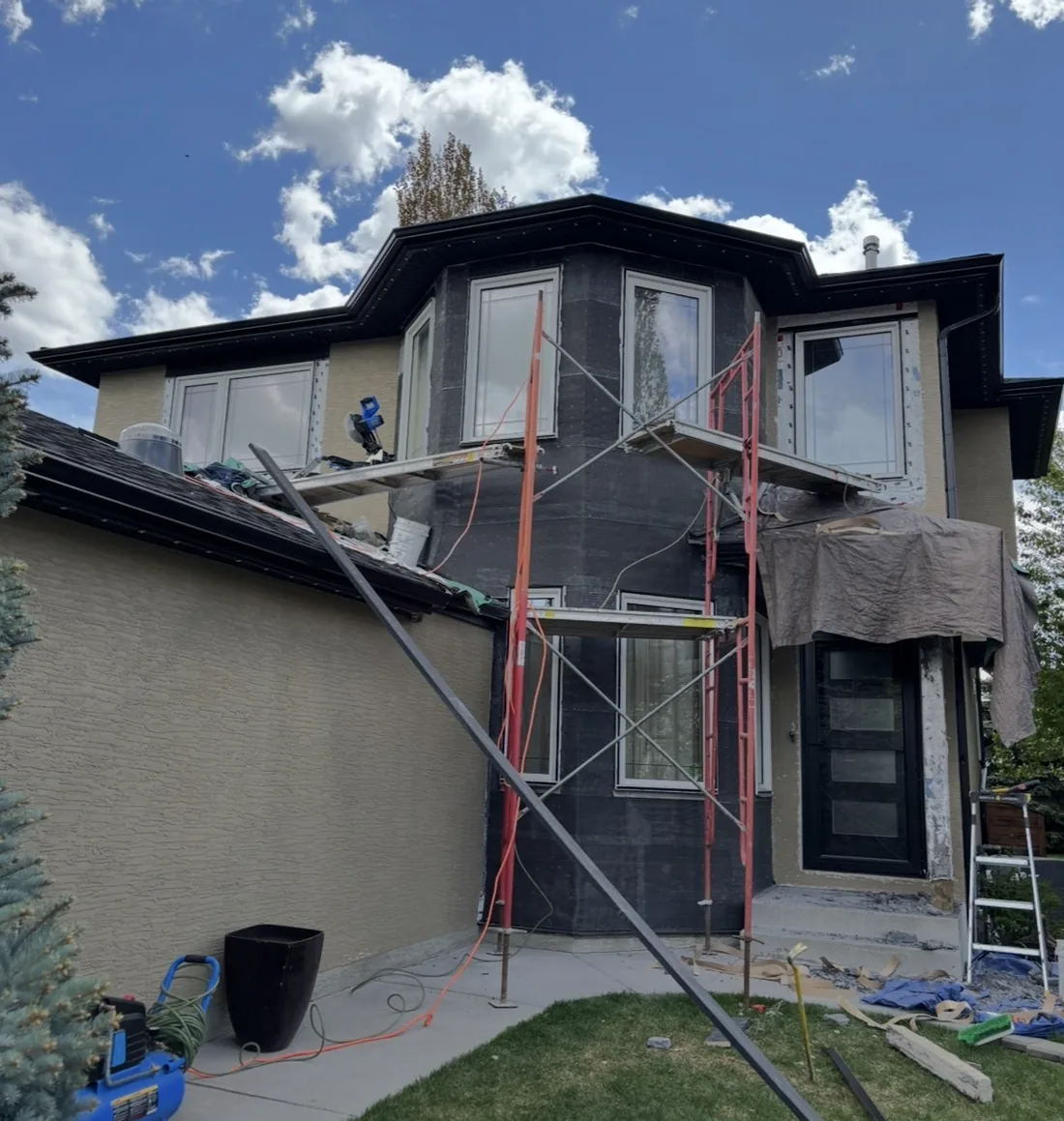 Calgary two-storey entry turret before stone cladding — bay wrapped in black weather membrane with scaffolding up, rest of the stucco finished