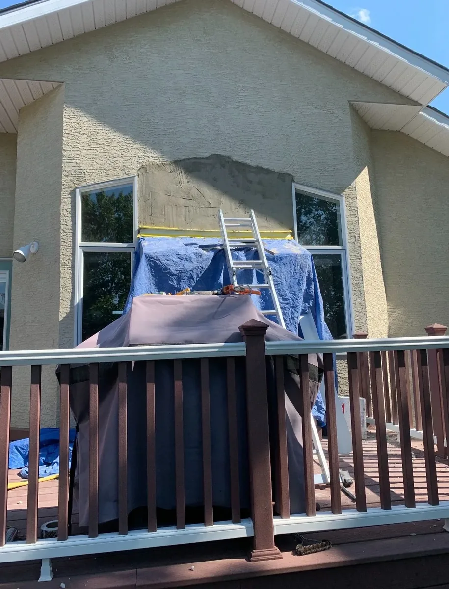 Calgary stucco wall leak repair before — failed flashing and water damage above window, work in progress with tarp and ladder