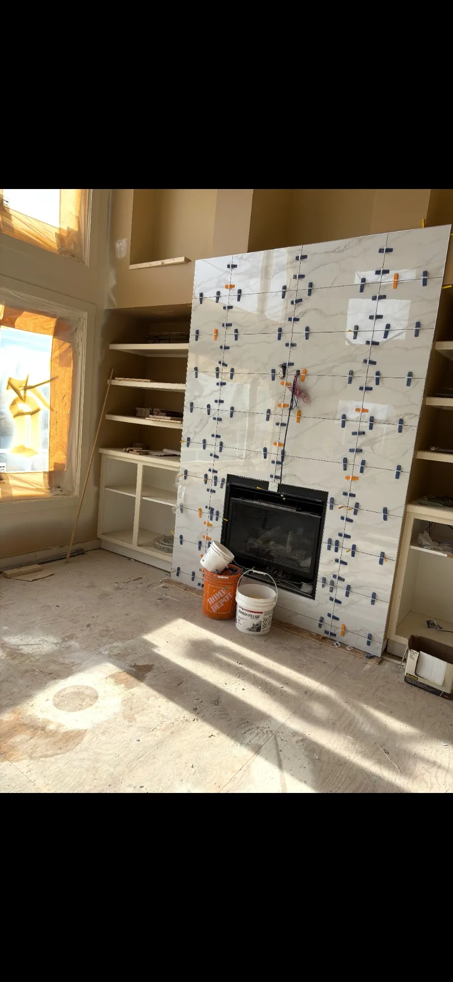 Calgary tile fireplace mid-install — large-format marble-look porcelain panel with lippage clips covering the surface, fireplace insert opening cut, buckets and tools in place