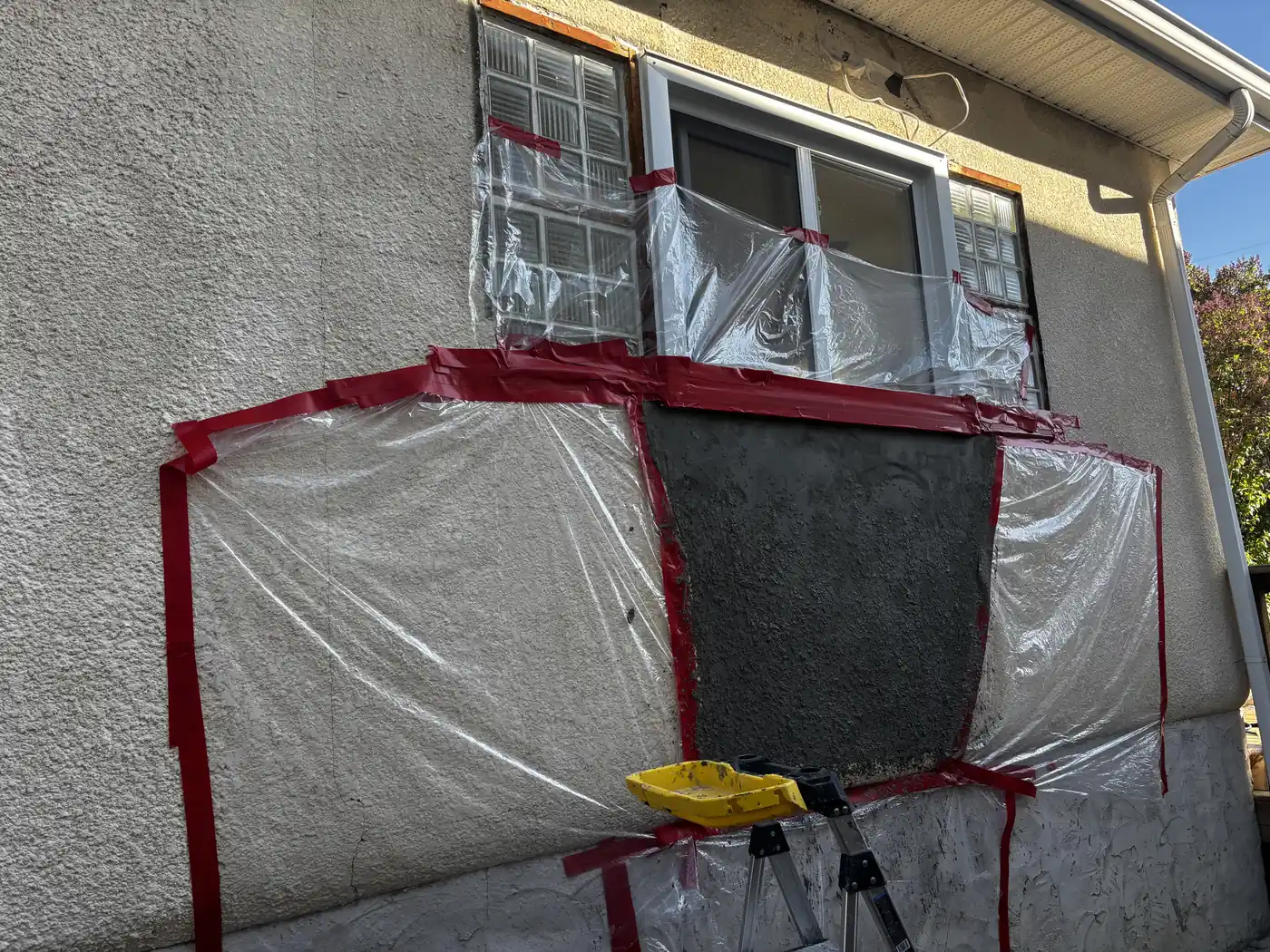 Calgary stucco leak repair below a window in progress, damaged section cut out and fresh base coat applied, windows masked off